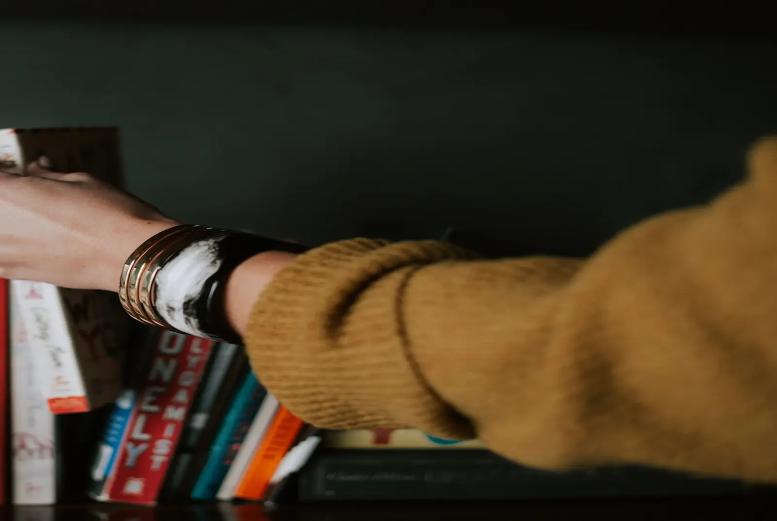 A hand in an ochre sweater pulling a title off a shelf in a dim room